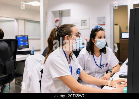Apothekerinnen, die Masken tragen, diskutieren über den Computer, während sie sitzen Krankenhaus Stockfoto