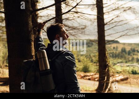 Nachdenklicher männlicher Wanderer mit Rucksack, der am Baumstamm steht Wald Stockfoto