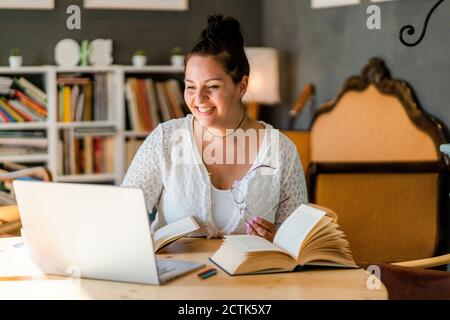 Lächelnde junge Frau studiert über Laptop an Büchern auf dem Tisch Im Café Stockfoto