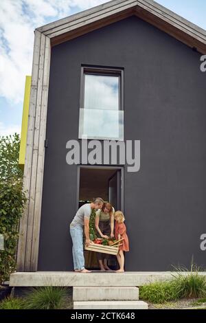Eltern mit Tochter, die Lebensmittel in der Kiste tragen, während sie stehen Eingang des winzigen Hauses Stockfoto