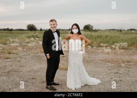 Lächelnde Braut mit Bräutigam trägt schützende Gesichtsmaske beim Blick Unterwegs im Feld während COVID-19 Stockfoto