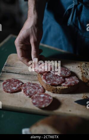 Hand des jungen Mannes, der Salami-Sandwich auf Schneidebrett vorbereitet Am Tisch Stockfoto