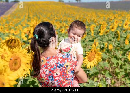 Mutter trägt fröhliche Tochter im Sonnenblumenfeld im Sommer Stockfoto