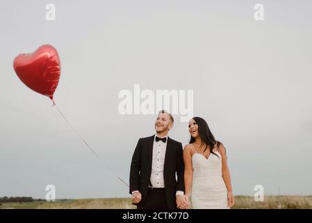 Glückliche Braut und Bräutigam mit Herz-Form Ballon gegen Himmel Stockfoto