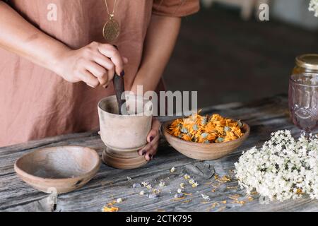 Mittelteil der Frau Zerkleinern Blumen und Kräuter in Mörtel mit Stößel auf Holztisch Stockfoto