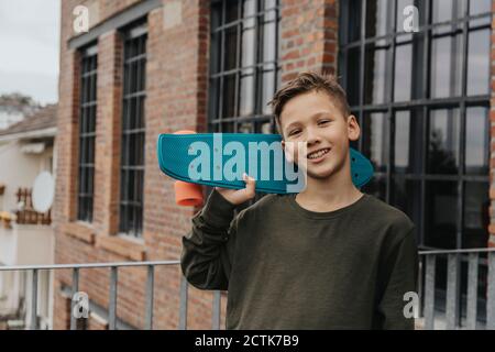 Lächelnder Junge posiert mit blauem Skateboard, während er gegen Gebäude steht Stockfoto