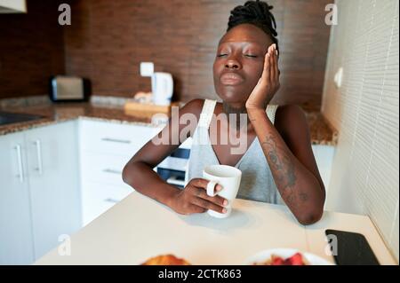 Junge Frau mit geschlossenen Augen sitzt mit Kaffeebecher an Tisch in der Küche Stockfoto