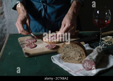 Mittelteil des jungen Mannes, der Salami-Sandwich auf Schneidebrett vorbereitet Am Tisch Stockfoto