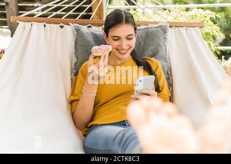 Lächelnd schöne Frau hält Donut, während mit Handy auf Hängematte im Hof Stockfoto