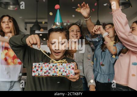 Neugierig Geburtstag junge Eröffnung Geschenk, während Freunde tanzen im Hintergrund Stockfoto