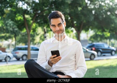 Selbstbewusster Geschäftsmann mit Smartphone, während er gegen Bäume sitzt Stockfoto