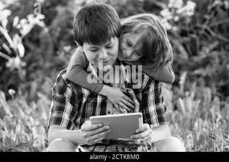 Teen junge mit seiner jüngeren Schwester sitzen in einem Park und mit dem Tablet. Schwarzweiß-Fotografie. Stockfoto