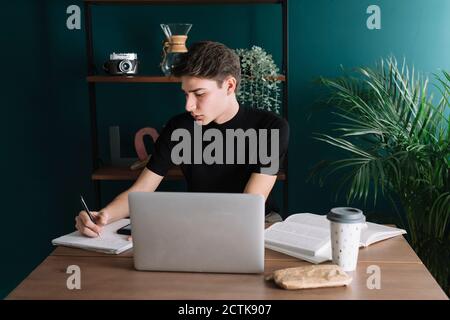 Hübscher junger Mann, der im Buch schreibt, während er mit Laptop sitzt Am Tisch Stockfoto