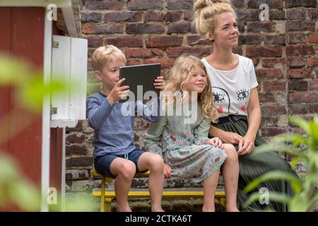 Junge fotografieren, während Schwester und Mutter gegen Backstein sitzen Wand Stockfoto