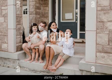 Lächelnde Familie mit Kindern, die während des Sitzens eine schützende Gesichtsmaske tragen Auf Stufen außerhalb des Hauses Stockfoto