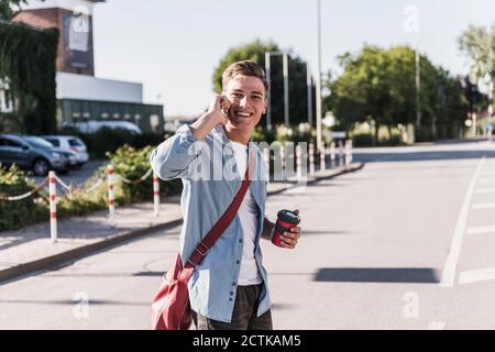 Lächelnder junger Mann, der auf dem Handy telefoniert, während er die Straße überquert In der Stadt Stockfoto