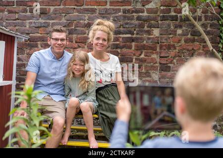 Junge fotografieren Familie sitzen an Backsteinwand durch digitale Tablet Im Hinterhof Stockfoto