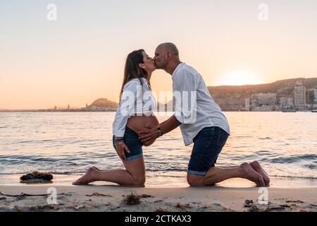Reifen Mann küssen schwangere Frau beim Knien gegen Strand während Sonnenuntergang Stockfoto