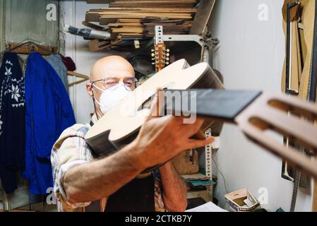 Handwerker mit Gesichtsmaske, die Gitarre beim Stehen in der Werkstatt untersucht Stockfoto