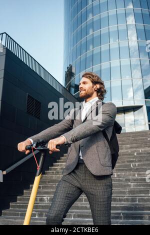 Handsome männlichen professionellen Blick weg, während mit elektrischen Push stehen Roller gegen Bürogebäude Stockfoto