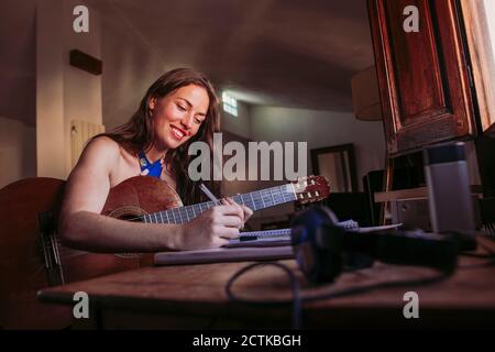 Lächelnde junge Frau, die im Buch schreibt, während sie Gitarre übt Zu Hause Stockfoto