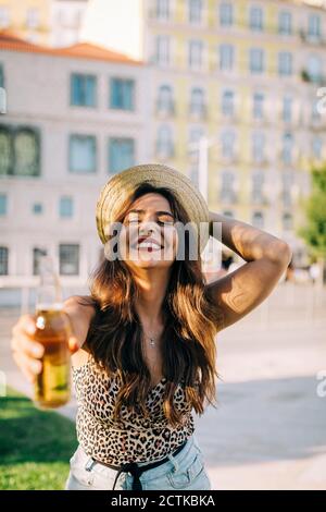Fröhliche junge Frau trägt Hut hält Bierflasche im Stehen In der Stadt Stockfoto