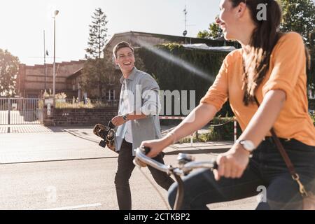 Fröhlicher junger Mann, der Freundin beim Fahrradfahren auf der Straße ansieht In der Stadt während des sonnigen Tages Stockfoto