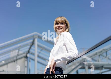 Lächelnde Unternehmerin, die wegschaut, während sie mit dem Geländer dasteht Blauer Himmel Stockfoto