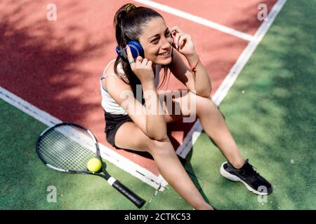 Lächelnde Tennisspielerin, die Musik hört, während sie auf dem Boden sitzt Vor Gericht Stockfoto