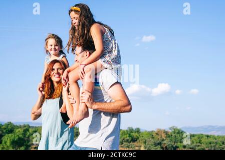 Glückliche Eltern tragen Töchter auf Schultern gegen den Himmel bei Sonnenschein Tag Stockfoto