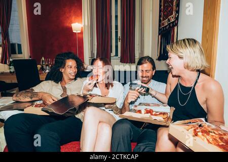 Glückliche Freunde essen Pizza, während sie auf dem Sofa im Wohnzimmer sitzen Zimmer Stockfoto