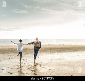 Fröhliches reifes Paar, das Hände hält, während es am Strand gegen springt wolkiger Himmel Stockfoto