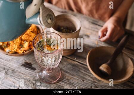 Teekanne gießt heißes Wasser in Kräuterteeglas auf den Tisch Stockfoto