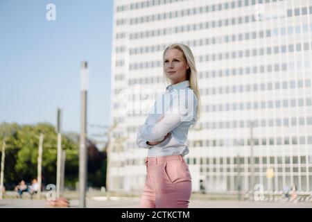 Nachdenkliche blonde professionelle Frau schaut weg, während sie mit Armen steht Gegen Gebäude im Finanzviertel gekreuzt Stockfoto