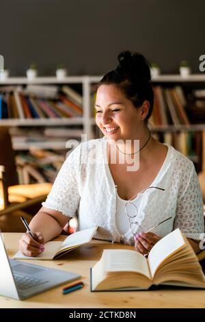 Lächelnde junge Frau, die im Buch schreibt, während sie über einen Laptop studiert Auf dem Tisch im Café Stockfoto