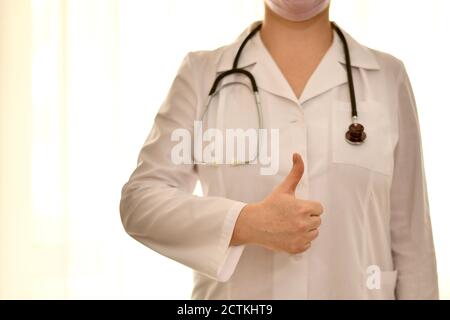 Arzt in weißer Robe mit Stethoskop zeigt Handgesten, ein gutes Symbol, einen erhobenen Daumen. Stockfoto