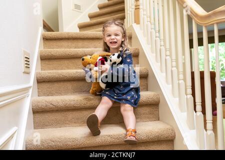 2.5-jähriges Mädchen, das auf der Treppe mit ausgestopften Tieren spielt und sich vorsichtig nach unten scooting (MR) Stockfoto