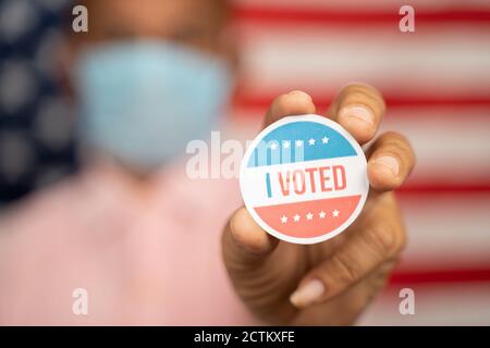 Nahaufnahme erschossen Mann in medizinische Maske zeigt ich gestimmt Aufkleber und setzen auf Shirt mit US-Flagge als Hintergrund - Konzept der US-Wahl. Stockfoto