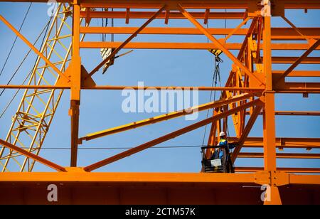 Kostanay/Kasachstan - Mai 14 2012: Bau eines neuen Fabrikgebäudes aus orangefarbenem Stahl. Arbeiter in aufgehängter Wiege und Trägerkran. Stockfoto