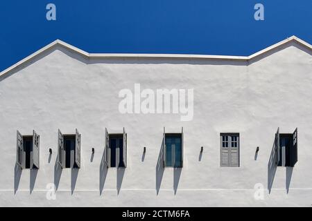 Eine weiße Steinwand mit einer Reihe von Fenstern mit offenen, geschlitzten, hölzernen Fensterläden, die nach unten Schatten wegen der hohen Mittagssonne schaffen. Im Gegensatz zu A Stockfoto