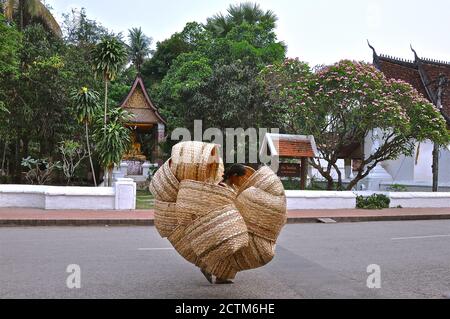 Ein Straßenhändler mit einem riesigen Stapel handgewebter Rattankörbe, der an einem buddhistischen Tempel in Laos vorbeiläuft. Stockfoto