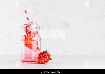 Erfrischendes kaltes Fruchtgetränk mit roter Erdbeere, Eis und Stroh im durchsichtigen Glas im eleganten weißen Interieur auf Holztisch, Kopierraum. Stockfoto