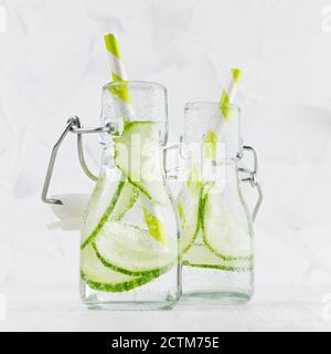 Natürliche Bio-Cocktails mit grüner Gurke, kohlensäurehaltiges Wasser, Scheiben und gestreiftem Stroh in Yoke-Flaschen auf hellweißem Holzbrett und Wand, quadratisch. Stockfoto