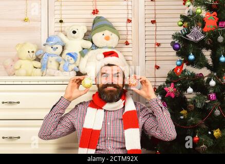 Guy schmückt Weihnachtsbaum. Weihnachtsmann mit glücklichem Gesicht auf Büro und Weihnachtsbaum Hintergrund. Feier- und Neujahrsdekor-Konzept. Mann mit Bart in Hut und Schal hält goldene Weihnachtskugeln Stockfoto