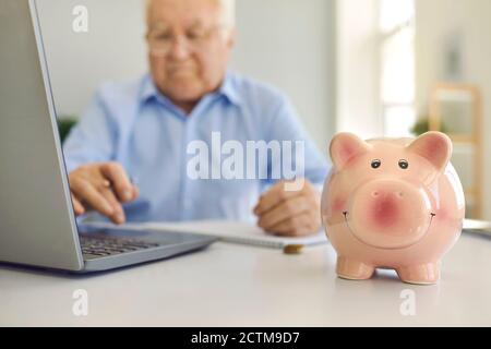 Sparschwein steht auf dem Schreibtisch mit verschwommenem älteren Mann Laptop, um Rechnungen online im Hintergrund zu bezahlen Stockfoto