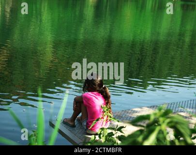 Nachdenkliches kleines Mädchen von hinten gesehen, auf einem Ponton am Flussufer sitzend, die Natur betrachtend Stockfoto