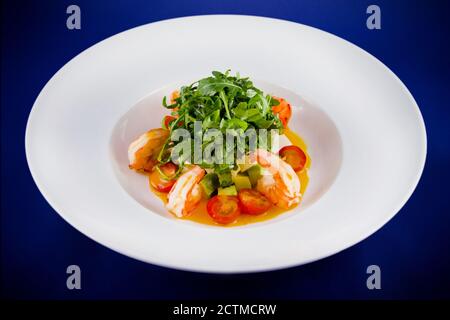 Salat mit Garnelen, Rucola, Tomaten, Avocado. Flach Lay Draufsicht auf einer weißen Platte auf einem blauen Hintergrund. Stockfoto