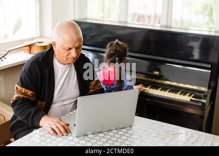 Kleines Mädchen spielt Klavier zusammen mit Großvater und lernt online auf einem Laptop Stockfoto