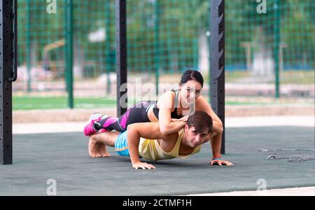 Junger athletischer Mann, der Liegestütze im Freien mit einer jungen Frau macht Sparschwein auf seinem Rücken in einem niedrigen Winkel Ansicht In einem Gesundheits- und Fitnesskonzept Stockfoto