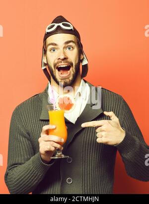 Schöner bärtiger Flieger Mann mit langem Bart auf lächelndem Gesicht hält Glas alkoholischen Cocktail in grau gestrickten Pullover mit Hut und Brille auf rotem Studio Hintergrund Stockfoto